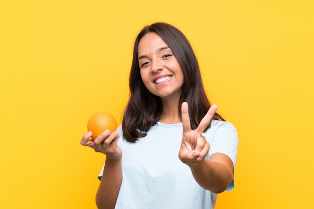 Jovem morena segurando uma laranja sorrindo e mostrando sinal de vitória