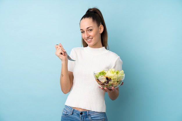 Jovem morena segurando uma salada sobre parede azul isolada