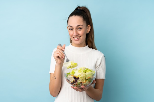 Jovem morena segurando uma salada sobre parede azul isolada