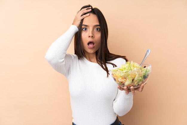 Jovem morena segurando uma salada sobre um fundo isolado com expressão de surpresa