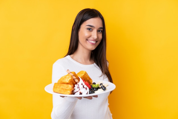 Jovem morena segurando waffles sobre parede isolada com expressão feliz