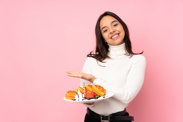 Jovem morena segurando waffles sobre parede rosa isolada, apresentando uma idéia enquanto olha sorrindo para