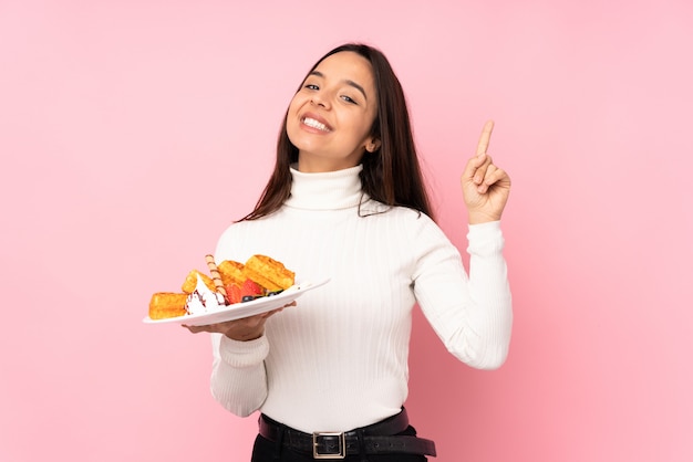 Jovem morena segurando waffles sobre parede rosa isolada, mostrando e levantando um dedo em sinal dos melhores