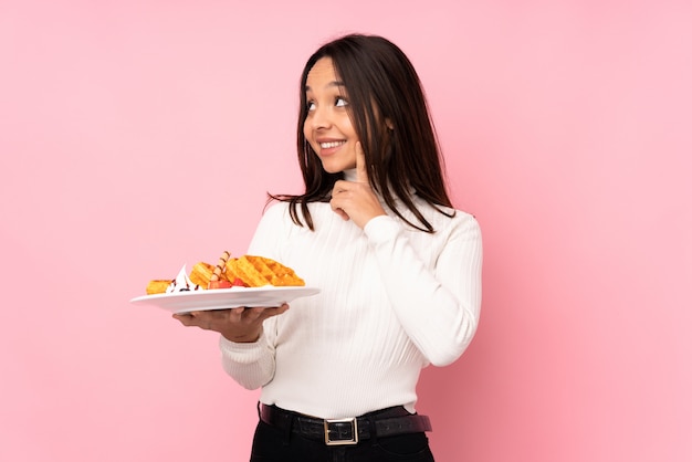 Jovem morena segurando waffles sobre parede rosa isolada, pensando uma idéia enquanto olha para cima