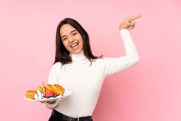 Jovem morena segurando waffles sobre um fundo rosa isolado apontando o dedo para as laterais e feliz