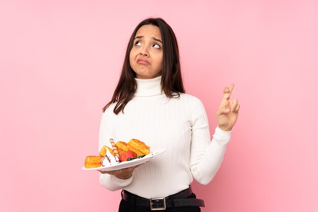 Jovem morena segurando waffles sobre um fundo rosa isolado com os dedos se cruzando e desejando o melhor