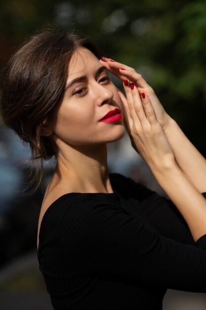 Jovem morena sensual com lábios vermelhos e manicure usando um vestido preto posando na rua com a luz do sol