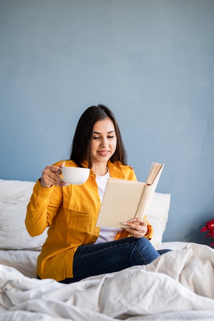 jovem morena sentada na cama comendo croissants e lendo um livro