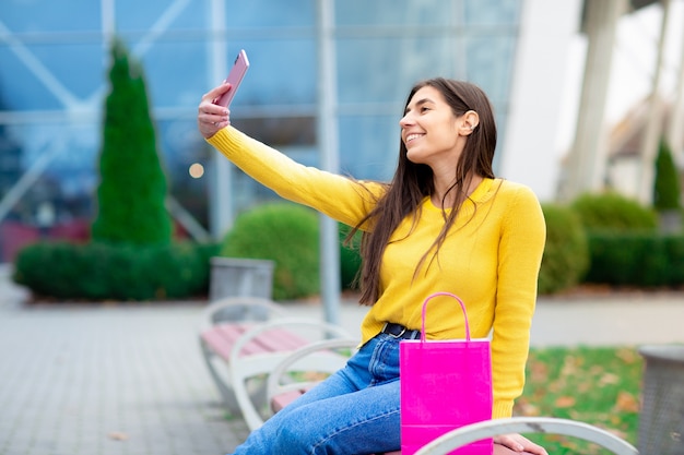 Jovem morena sentado ao ar livre no banco com sacolas rosa e fazendo selfies. mulher vestida de camisola amarela