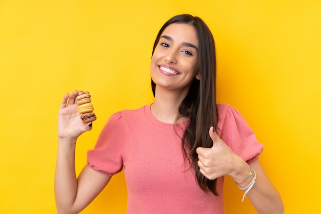 Jovem morena sobre parede amarela isolada segurando macarons franceses coloridos com polegares