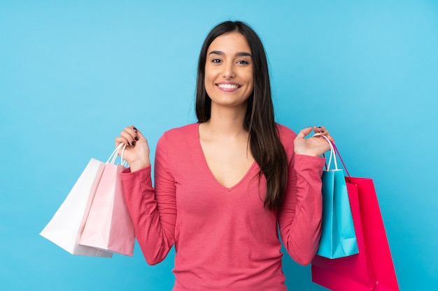 Jovem morena sobre parede azul isolada segurando sacolas de compras e sorrindo