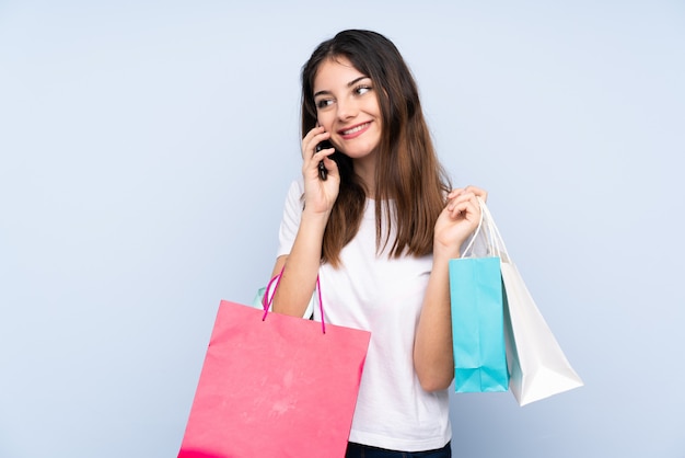 Jovem morena sobre parede azul segurando sacolas de compras e ligando com o celular