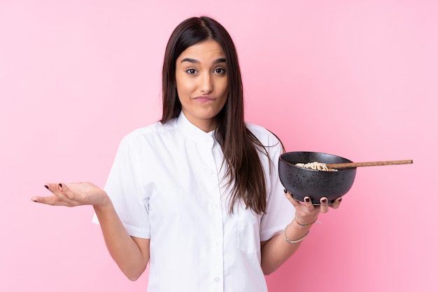 Jovem morena sobre parede rosa isolada, fazendo dúvidas gesto enquanto levanta os ombros enquanto segura uma tigela de macarrão com pauzinhos