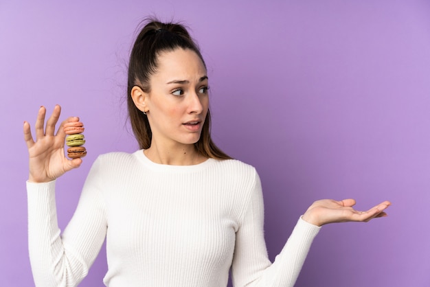 Foto jovem morena sobre parede roxa isolada segurando macarons franceses coloridos e com expressão de surpresa