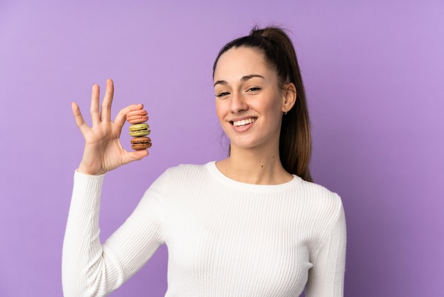 Jovem morena sobre parede roxa isolada segurando macarons franceses coloridos e sorrindo muito