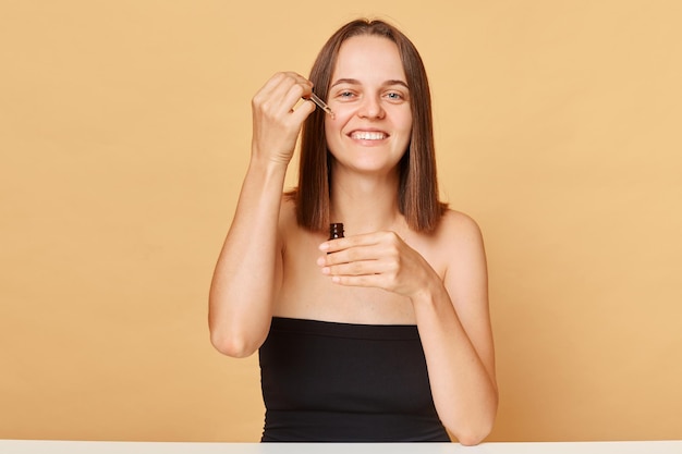 Foto jovem morena sorridente aplica essência vestindo top preto posando isolado sobre fundo bege, desfrutando de procedimentos de cuidados com a pele na manhã de fim de semana