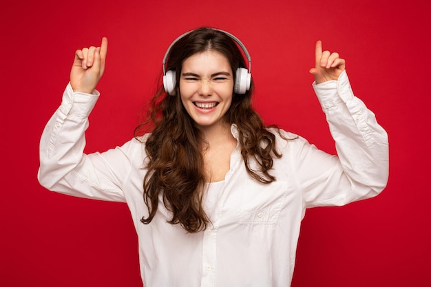 Jovem morena sorridente atraente feliz vestindo camisa branca e óculos ópticos isolados sobre fundo vermelho usando fones de ouvido bluetooth sem fio brancos, ouvindo música e dançando