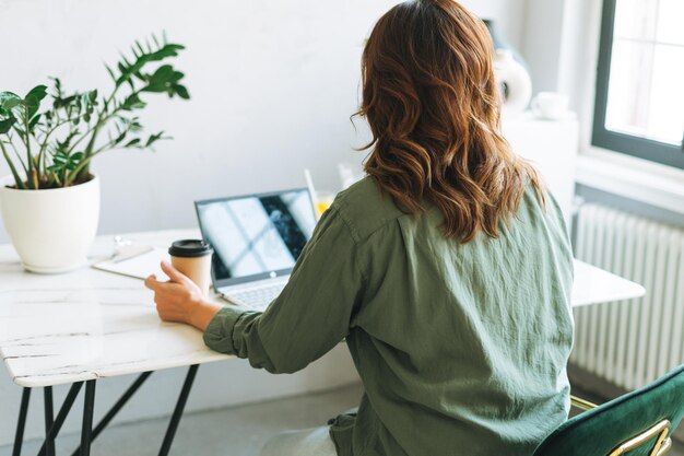 Jovem morena sorridente de tamanho maior trabalhando no laptop na mesa com a planta da casa na vista de trás do escritório moderno e brilhante