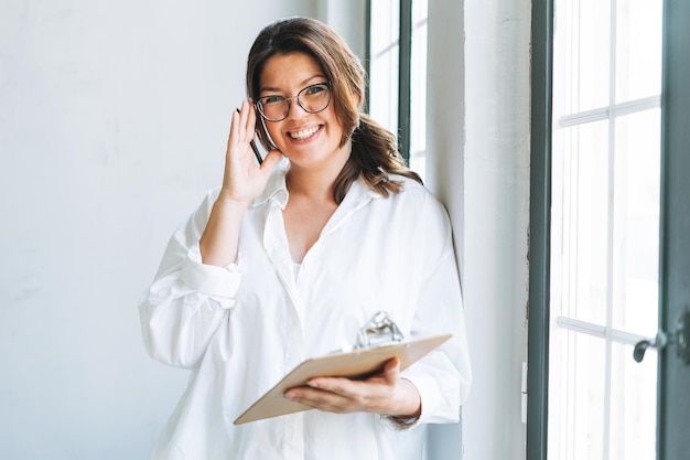 Foto jovem morena sorridente mais tamanho na camisa branca perto da janela com caneta e documentos nas mãos no escritório moderno brilhante