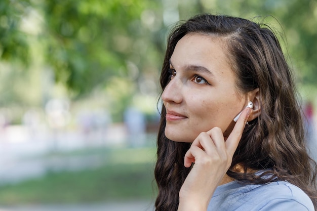 Jovem morena tocando fones de ouvido TWS enquanto ouve música durante caminhada ao ar livre