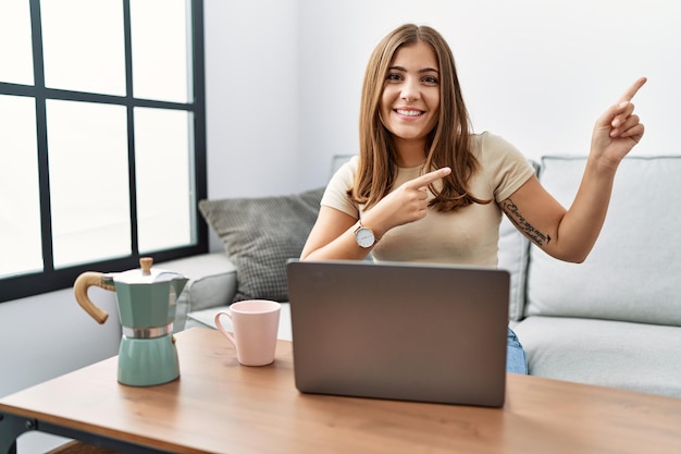 Jovem morena usando laptop em casa bebendo uma xícara de café sorrindo e olhando para a câmera apontando com as duas mãos e dedos para o lado