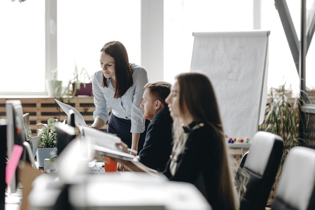 Jovem morena vestida com roupas de estilo office está sentada à mesa com os colegas e mantendo o notebook nas mãos em um escritório moderno e leve