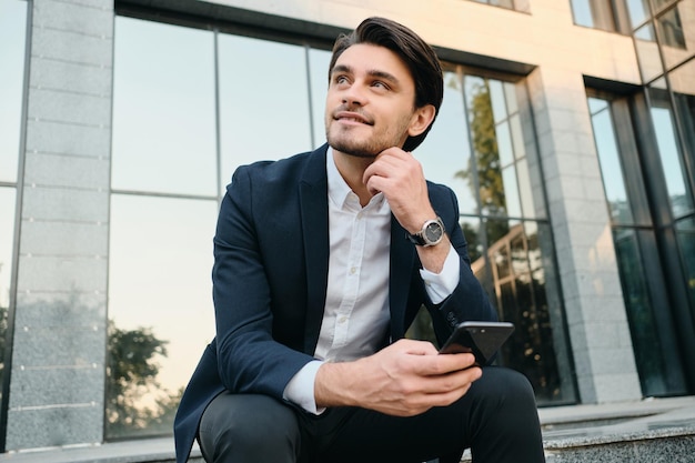 Jovem moreno barbudo sorridente bonito na camisa branca e terno clássico olhando alegremente de lado segurando o celular na mão enquanto está sentado na escada com vidro construindo no fundo