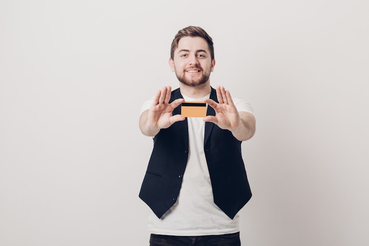 Foto jovem moreno bonito com uma barba em uma camisa branca e um colete preto segurando um cartão de crédito de plástico e sorrindo. concentre-se no cartão. luz suave