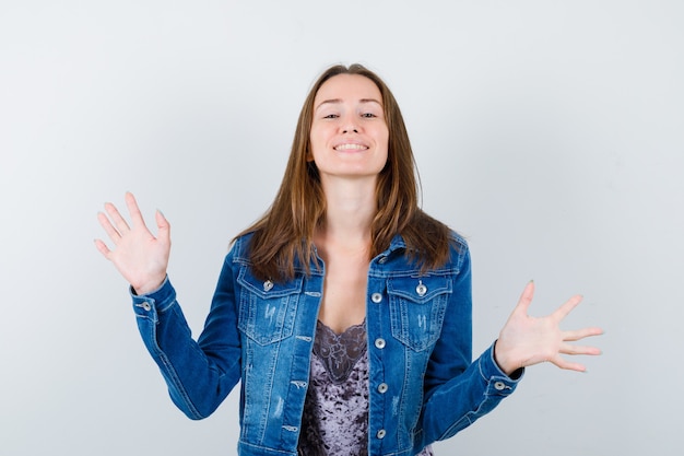 Foto jovem, mostrando as palmas das mãos em gesto de rendição na blusa, jaqueta jeans e olhando feliz, vista frontal.