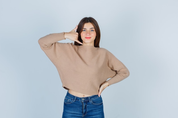 Jovem, mostrando o gesto do telefone em um suéter bege, jeans e um olhar alegre. vista frontal.