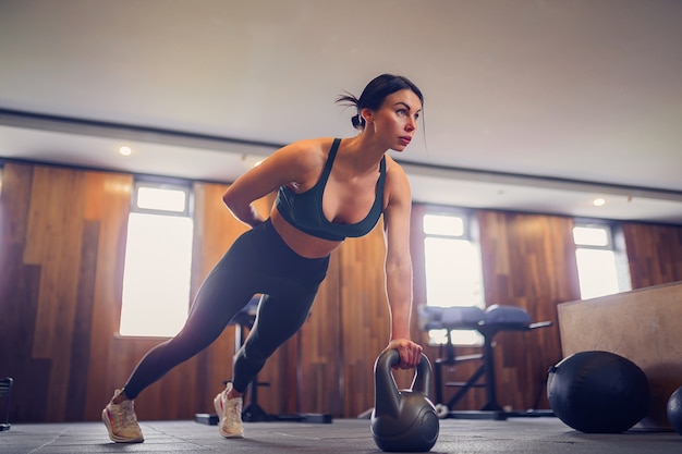 Jovem motivado fazendo exercício de prancha usando kettlebells com uma mão no ginásio