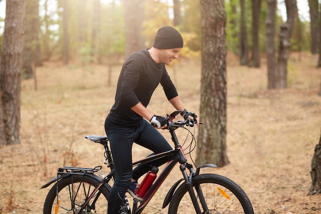 Jovem motociclista desportivo, andar de bicicleta na paisagem inspiradora floresta. homem de bicicleta mtb no caminho de trilha de enduro, cobrindo a distância atribuída, vestindo roupas esportivas pretas e boné, aproveitando a recreação ativa.