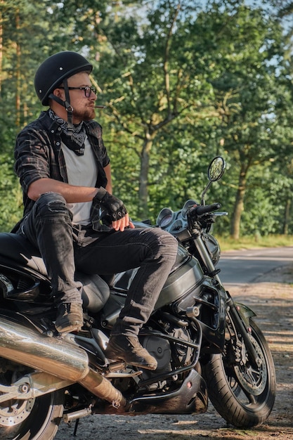Jovem motociclista masculino no capacete viaja de moto sozinho parado e fumando ao lado de uma floresta