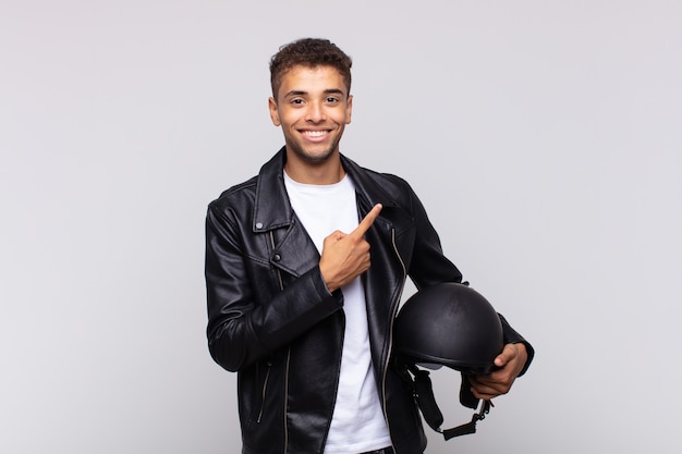 Jovem motociclista sorrindo alegremente, sentindo-se feliz e apontando para o lado e para cima, mostrando o objeto no espaço da cópia