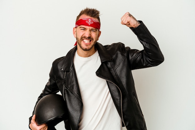 Jovem motociclista tatuado homem caucasiano segurando um capacete isolado no fundo branco, levantando o punho após uma vitória, o conceito de vencedor.