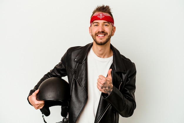 Jovem motoqueiro tatuado homem caucasiano segurando um capacete isolado no fundo branco, sorrindo e levantando o polegar