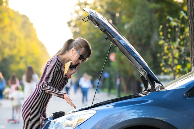 Jovem motorista chateada falando ao celular perto de um carro quebrado com o capô aberto, esperando por ajuda, tendo problemas com seu veículo em uma rua da cidade