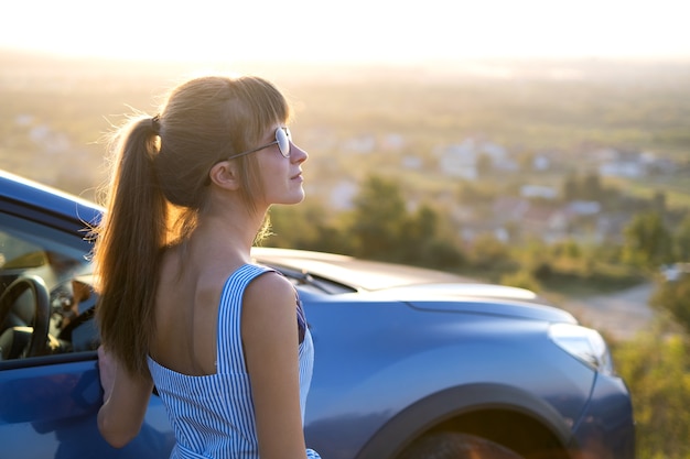 Foto jovem motorista do sexo feminino descansando perto de seu carro, aproveitando a noite quente de verão. conceito de viagens e fuga.