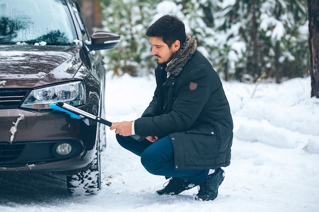 Jovem motorista, um homem, limpa os faróis de um carro da neve no inverno