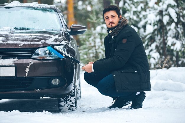 Jovem motorista, um homem, limpa os faróis de um carro da neve no inverno