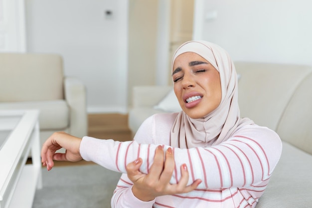 Jovem muçulmana árabe com hijab está sentada em casa no sofá e tocando o cotovelo enquanto sofre de dor no cotovelo