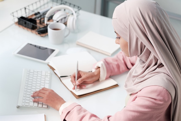 Jovem muçulmana contemporânea usando hijab e roupa casual elegante, sentada à mesa em frente ao monitor do computador no escritório e fazendo anotações