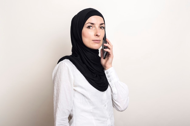 Jovem muçulmana em hijab falando ao telefone e olhando para a câmera