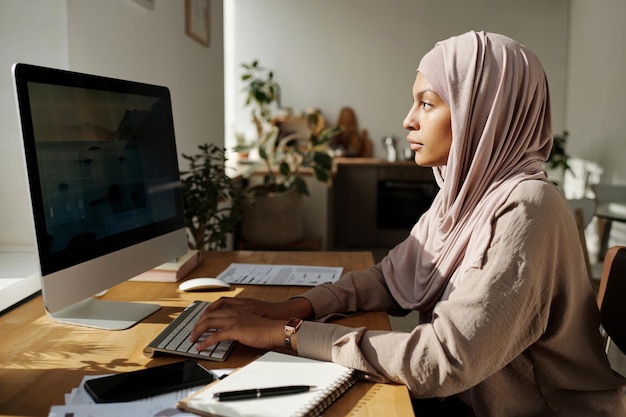 Jovem muçulmana séria de hijab digitando no teclado do computador e olhando para a tela