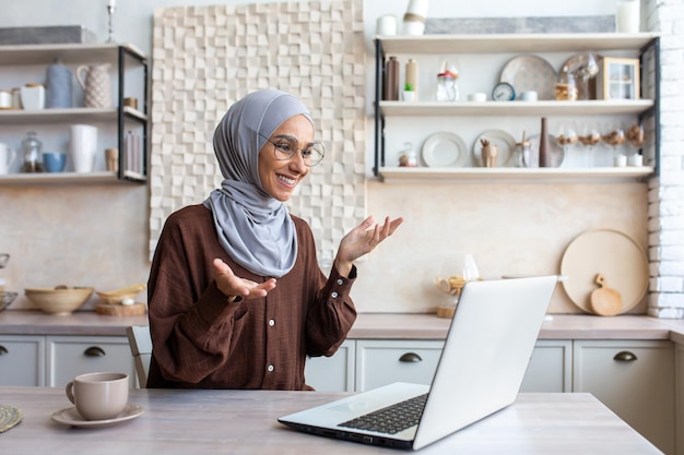 Jovem muçulmana sorridente em hijab sentada na cozinha em casa e conversando em videochamada via laptop