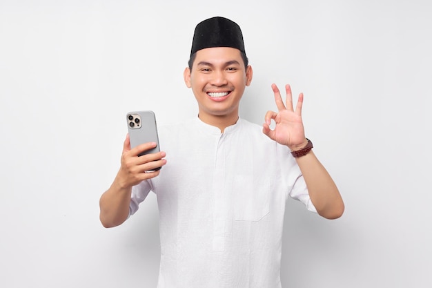 Jovem muçulmano asiático sorridente segurando smartphone e gesticulando sinal de ok isolado no fundo branco Celebração do conceito de estilo de vida do Islã religioso Ramadã e Ied Mubarak