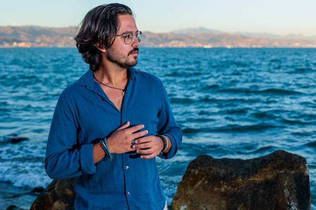 jovem muito atraente com camisa azul e calças brancas posando na praia com o mar ao fundo