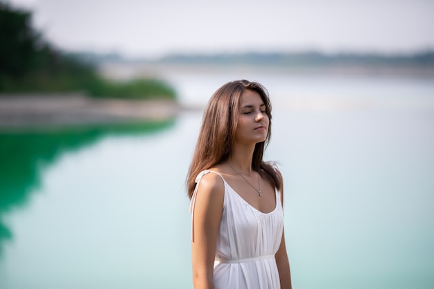 Jovem muito bonita com cabelos longos em um vestido branco à beira do lago.