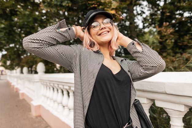 Jovem muito sorridente com um lindo rosto emocional em óculos da moda em roupas elegantes, desfrutando de um passeio no parque