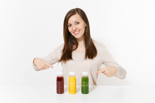 Jovem mulher à mesa com smoothies de desintoxicação verdes, vermelhos e amarelos em garrafas isoladas no fundo branco. Nutrição adequada, bebida vegetariana, estilo de vida saudável, conceito de dieta. Área com espaço de cópia.